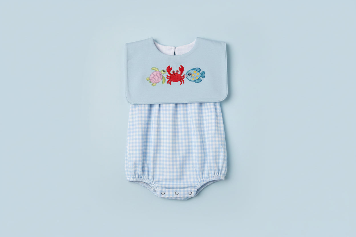 Embroidered bib with sea creature designs on a light blue background, held over a checkered garment.
