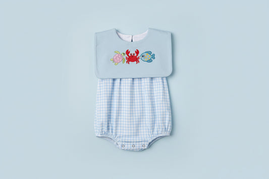 Embroidered bib with sea creature designs on a light blue background, held over a checkered garment.