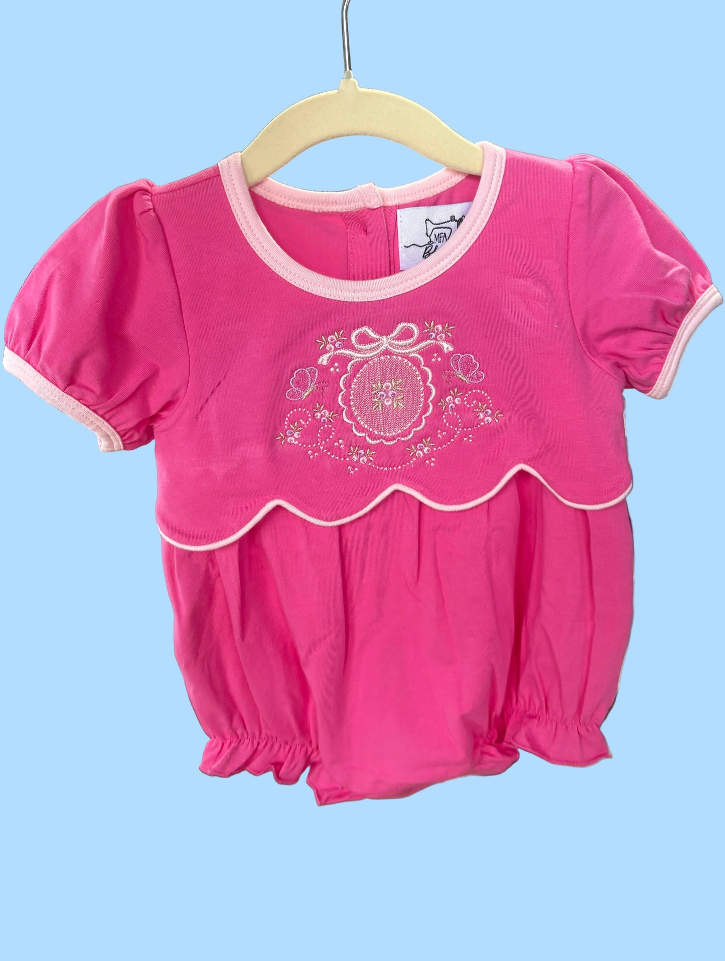 Pink baby outfit with white trim on a hanger against a blurred indoor background